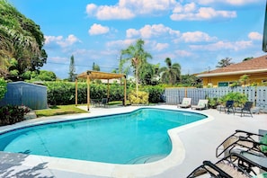 A swimming pool with a wooden pergola, lounge chairs, and a fenced yard with palm trees.