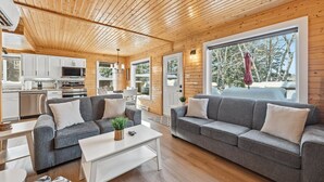 A modern cabin interior with wooden ceilings, a kitchen, a living area with two grey sofas, and large windows offering a view of trees. - Lakeside Retreat with Hot Tub (Windsor)