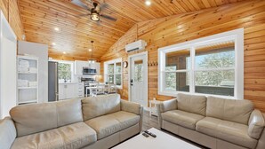 A cozy cabin interior with two beige leather sofas, a white coffee table, a kitchen area, and large windows offering a view of the outdoors. - Treeside Retreat with Hot tub (Windsor)