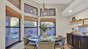 A dining area with a round table, six chairs, a chandelier, and large windows with blinds.