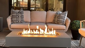 A beige sofa with patterned cushions, a rectangular fire feature, and a view of trees through large windows.