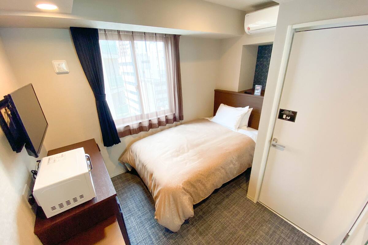 A hotel room with a bed, a desk with a computer, a window with curtains, and an air conditioning unit.