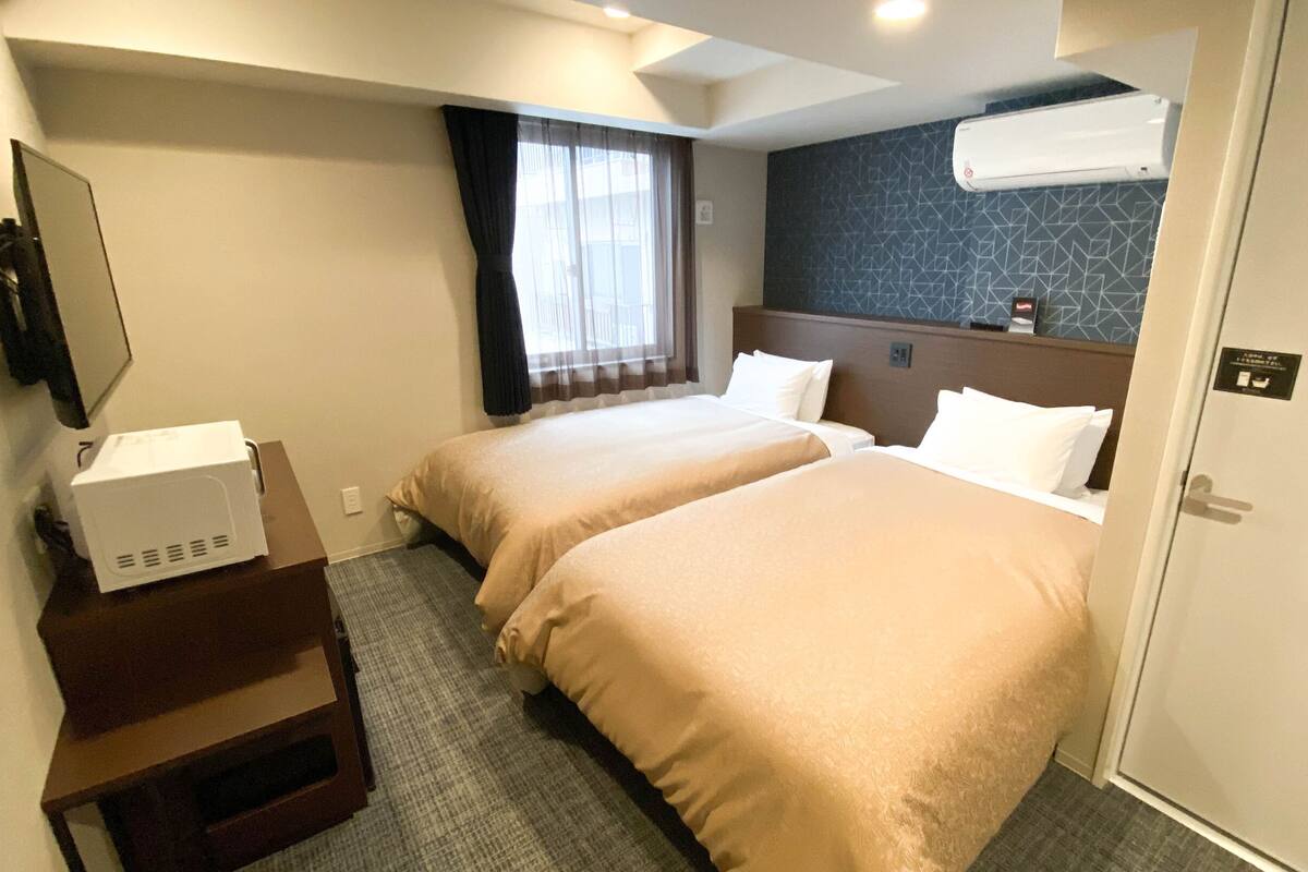 A hotel room with two beds, a desk with a computer, a window with curtains, an air conditioning unit, and a door with a sign.