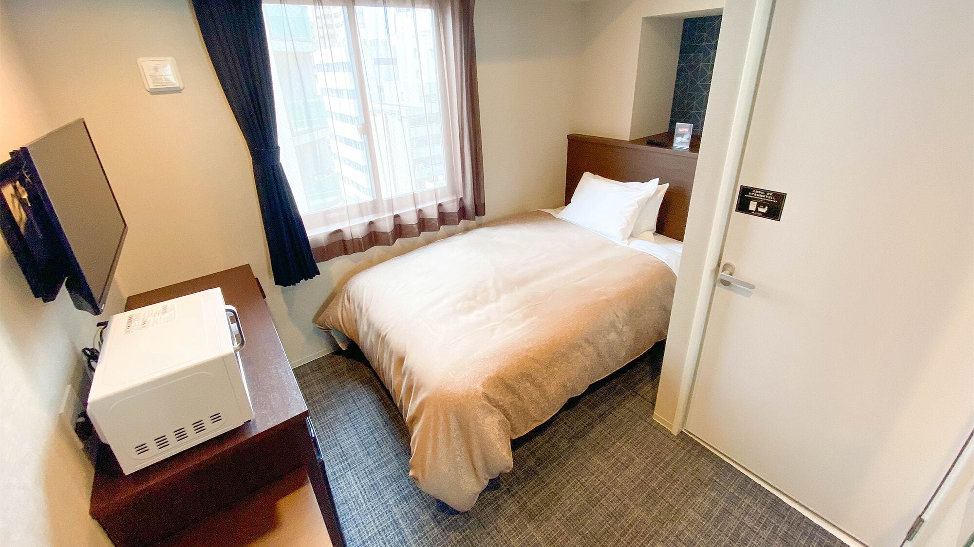 A hotel room with a bed, a desk with a white appliance, a window with curtains, and a door with a sign.