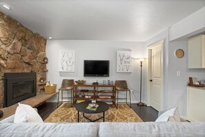 A living room with a stone wall, a fireplace, a flat-screen TV, a coffee table, two armchairs, and a bookshelf.