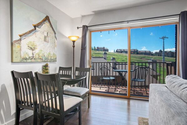 A room with a dining area, a sofa, and a balcony with a view of houses and greenery.