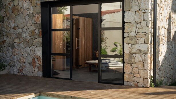 Una casa moderna con fachada de piedra, una terraza de madera y una puerta de cristal que da a un interior con un banco de madera y una planta.