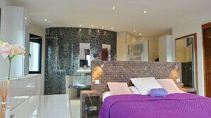 Une chambre à coucher moderne avec un grand lit, une salle de bain dotée d’une douche en verre et une coiffeuse blanche.