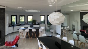 Une cuisine moderne dotée d’un îlot central, de grandes fenêtres et d’un coin repas avec une table noire et des chaises blanches.