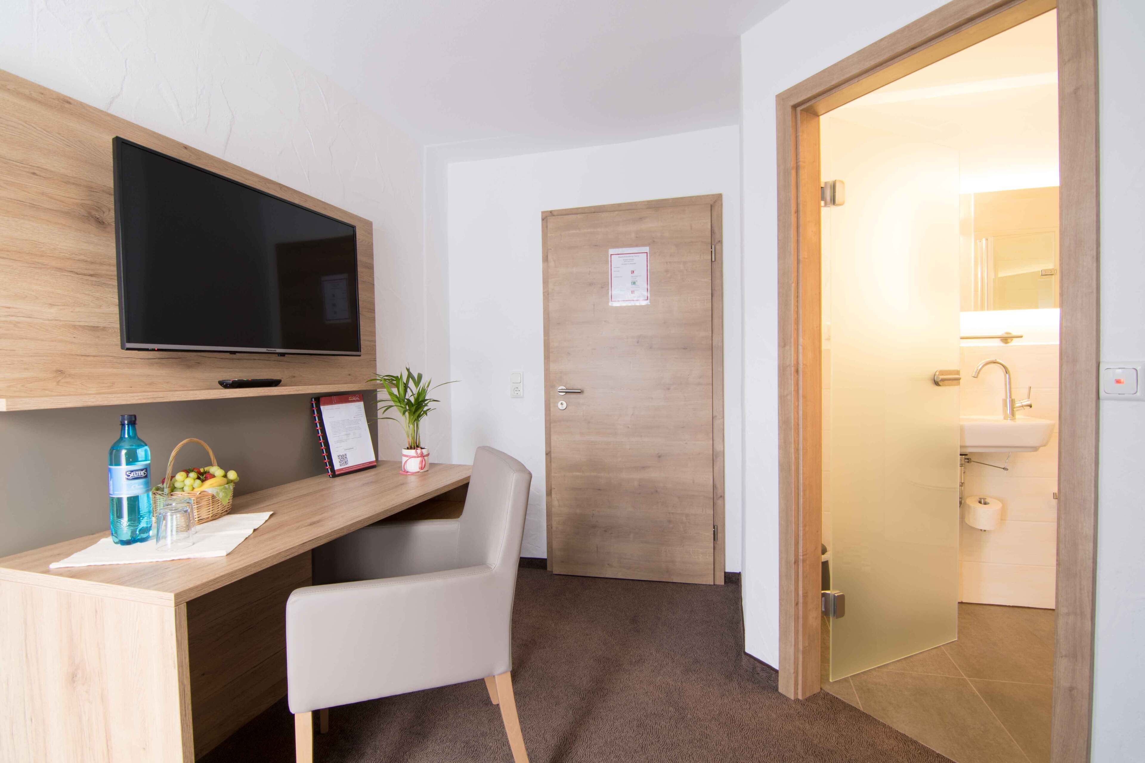 Habitación de hotel con escritorio de madera, televisor de pantalla plana, una silla, planta en maceta y baño con lavamanos e inodoro visibles a través de una puerta abierta.