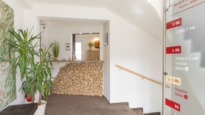 A hotel reception area with a staircase leading to multiple floors, potted plants, a stone wall, and a glass door with room numbers and directions. - Pension Klein (Bad Laasphe)