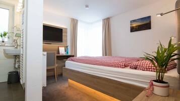 Ett hotellrum med en säng, ett skrivbord, en tv, ett fönster med gardiner, en växt och en tavla på väggen.
