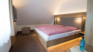 A bedroom with a wooden bed frame, a bedside table with a lamp, a small stool, and a TV mounted on the wall. - Pension Klein (Bad Laasphe)
