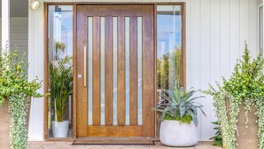 A wooden front door with vertical glass panels, flanked by potted plants on either side.