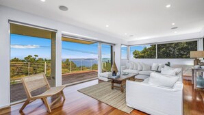 A spacious living room with a large white sofa, a wooden coffee table, and a wicker chair. There are large windows offering a view of the outdoors.