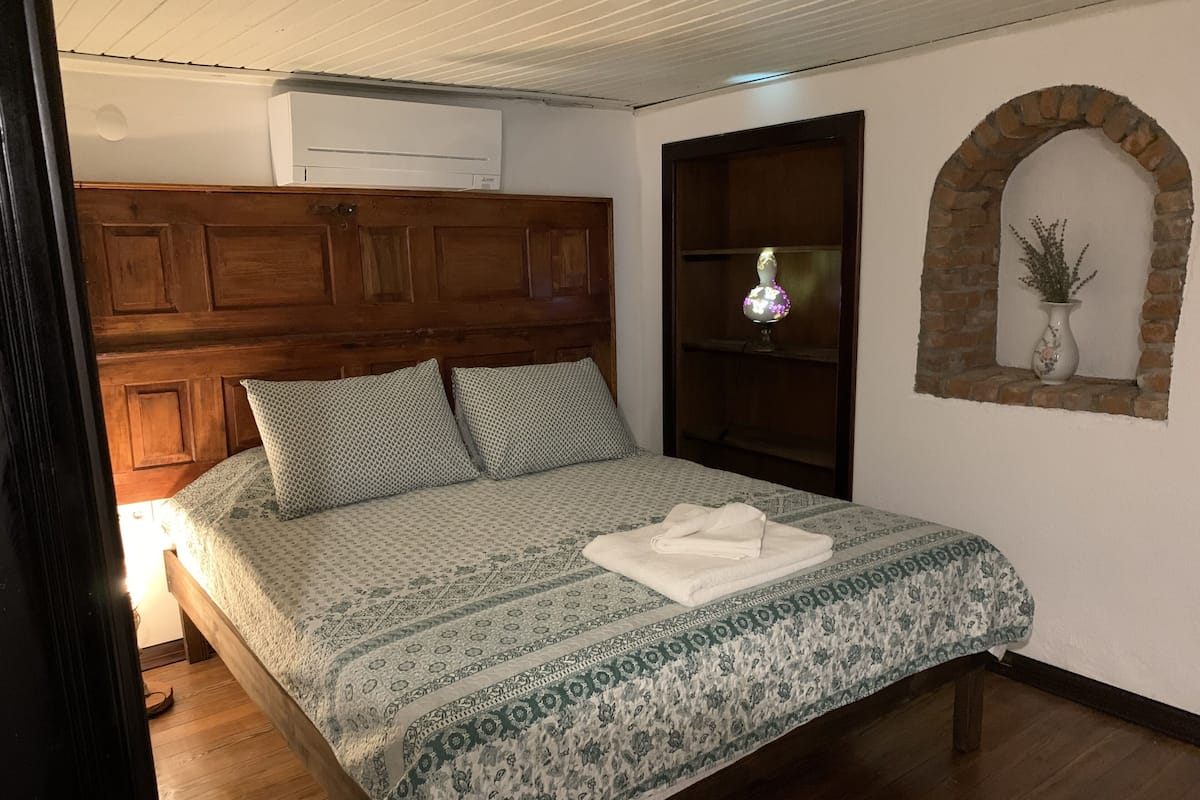 A bedroom with a wooden headboard, a bed with patterned bedding, a nightstand, a globe, and a brick archway with a vase.