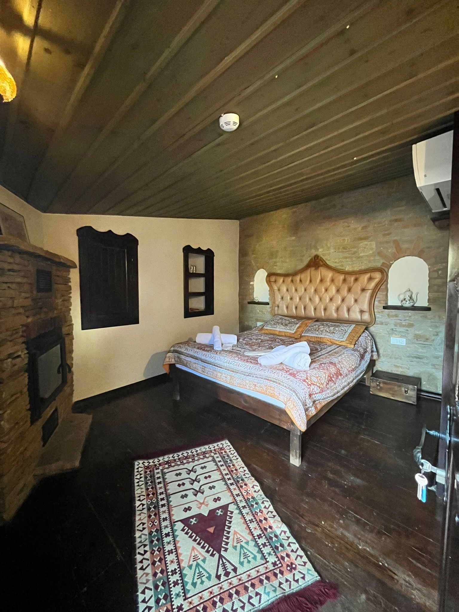 A bedroom with a wooden ceiling, a bed with a patterned bedspread, a decorative headboard, a stone wall, and a small window.