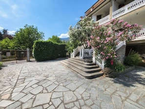 Une entrée dallée de pierres, avec un escalier menant à un balcon, le tout entouré de verdure et sous un ciel dégagé.