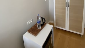 Ein Zimmer mit weißem Schreibtisch, einer Wasserflasche und einem Wasserkocher. Ein Kleiderschrank mit zwei Türen und eine weiße Wand mit Lichtschalter.