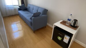 Ein Zimmer mit einem blauen Sofa, einem kleinen weißen Tisch mit Wasserflasche und Wasserkocher sowie einem Kühlschrank mit Energielabel A+.