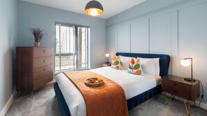 A bedroom with a bed, bedside table, lamp, dresser, window, and a potted plant. - Elliot Oliver - Cosy 2 Bedroom Town Centre Apartment (Cheltenham)