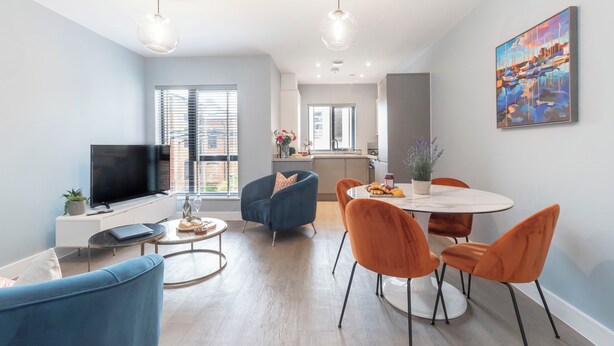 A modern living room with a flat-screen TV, blue and orange chairs, a dining table, and a kitchen in the background. - Elliot Oliver - Cosy 2 Bedroom Town Centre Apartment (Cheltenham)