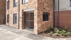 A brick building with a gated entrance, a small garden area with plants, and a concrete pathway. - Elliot Oliver - Cosy 2 Bedroom Town Centre Apartment (Cheltenham)