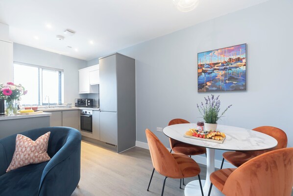 A modern kitchen with white cabinets, a dining area with a marble table, and a painting of a harbor scene.