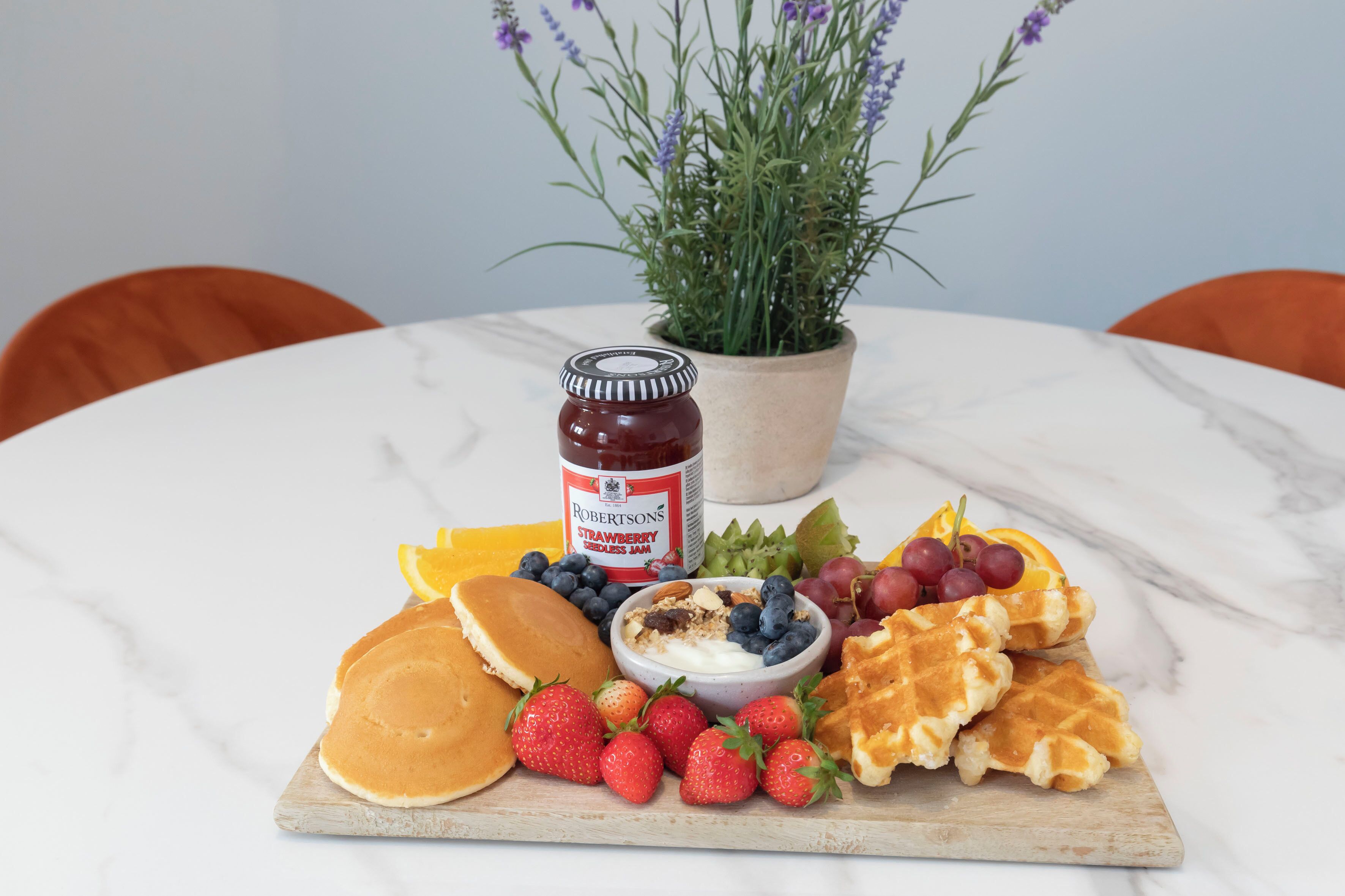 A breakfast platter with pancakes, waffles, strawberries, blueberries, grapes, and a jar of strawberry jam on a marble table.