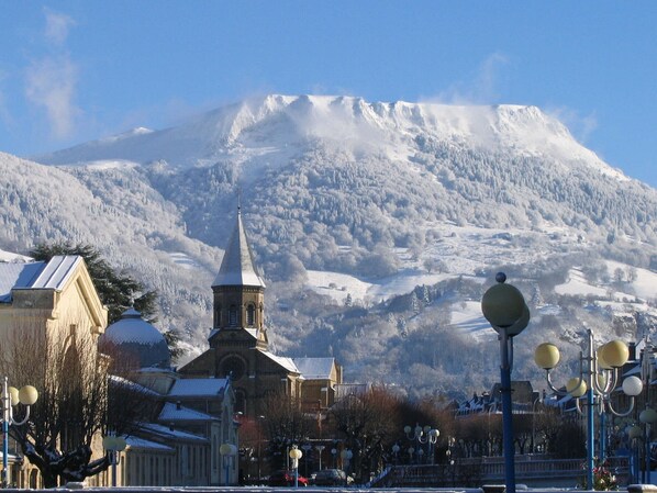 A town with a church featuring a tall steeple, surrounded by snow-covered buildings and a mountain in the background. - T3 Apartment near La Bourboule center with terrace (La Bourboule)