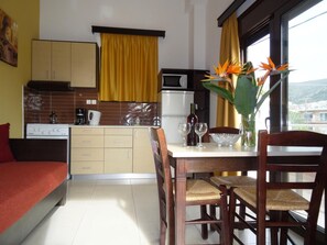 A compact kitchen with a dining area, featuring a dining table with chairs, a small refrigerator, a microwave, and a window with curtains. - Lito Apartments (Kantanos-Selino)