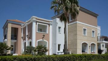 A multi-story building with balconies and arched windows, surrounded by greenery and palm trees.