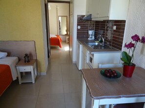 A compact hotel room with a bed, a small table with a fruit bowl, a nightstand, a kitchenette, and a bathroom visible in the background. - Lito Apartments (Kantanos-Selino)