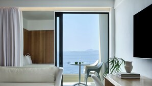 Ett modernt hotellrum med en säng, ett skrivbord, en stol, ett litet bord med två flaskor och utsikt över havet.
