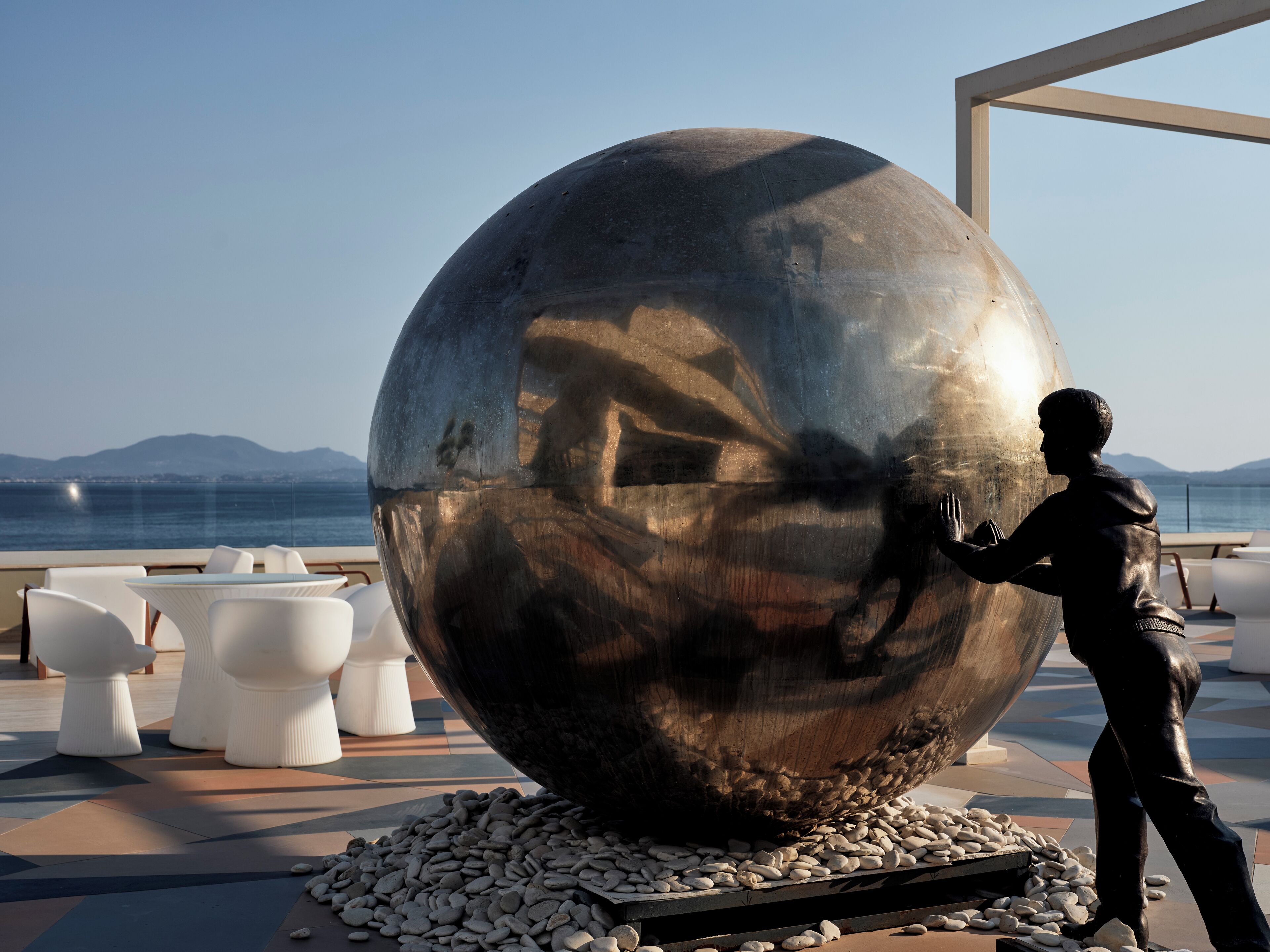 A large metallic sculpture of a man reaching out, surrounded by white chairs and a pebble-lined area, with a view of the sea and mountains in the background.