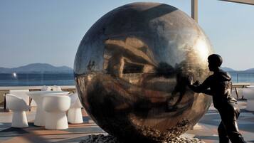 A large metallic sculpture of a man reaching out, surrounded by white chairs and a pebble-lined area, with a view of the sea and mountains in the background.