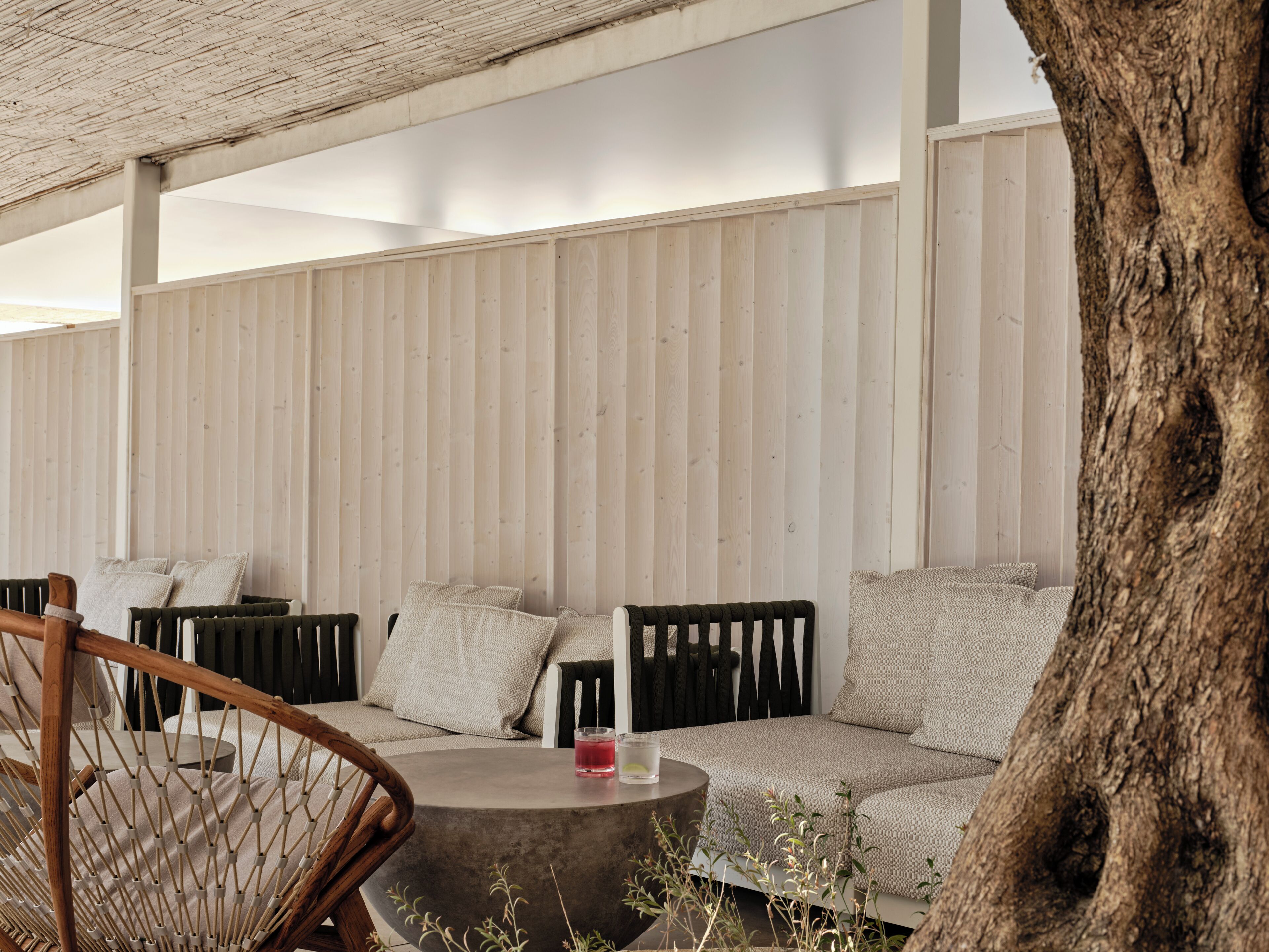 A covered outdoor seating area with a woven chair, a round table, and a cushioned sofa against a wooden wall.