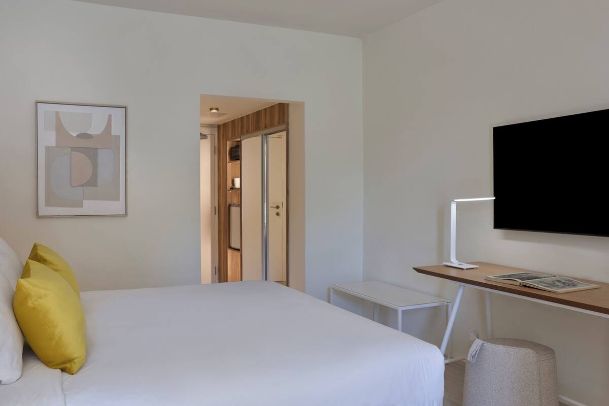 Une chambre d’hôtel avec un lit, un bureau équipé d’un ordinateur, une télévision et une œuvre d’art abstraite encadrée accrochée au mur.