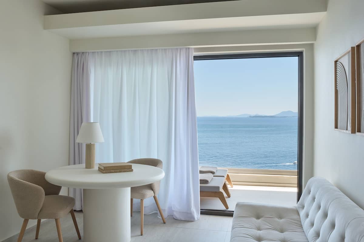 Une pièce moderne avec une table ronde blanche, deux chaises, un canapé blanc et une vue sur la mer grâce à de grandes fenêtres.