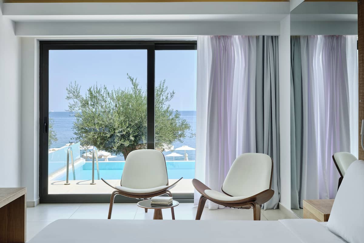 Une chambre d’hôtel moderne dotée d’une grande porte-fenêtre coulissante donnant sur un espace piscine, de deux chaises blanches, d’un lit et d’une table de chevet.