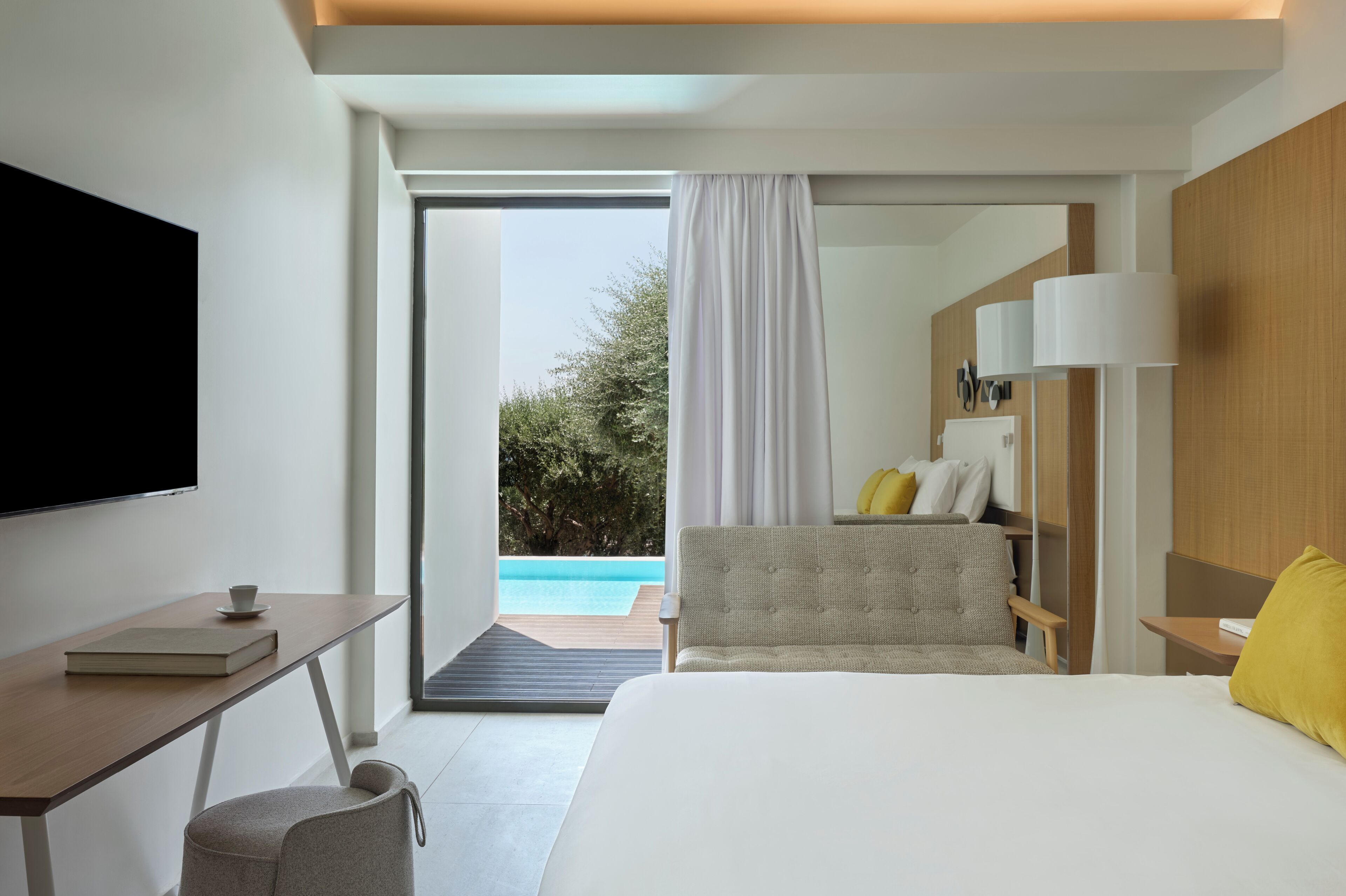A modern hotel room with a bed, a desk with a book and cup, a TV, and a view of a swimming pool.