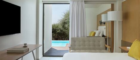 A modern hotel room with a bed, a desk with a book and cup, a TV, and a view of a swimming pool.
