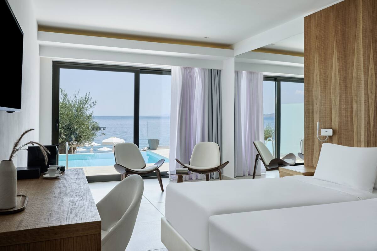 Une chambre d’hôtel avec un grand lit, deux fauteuils, un bureau agrémenté d’un vase et de tasses, et une vue sur la mer grâce à de grandes fenêtres.