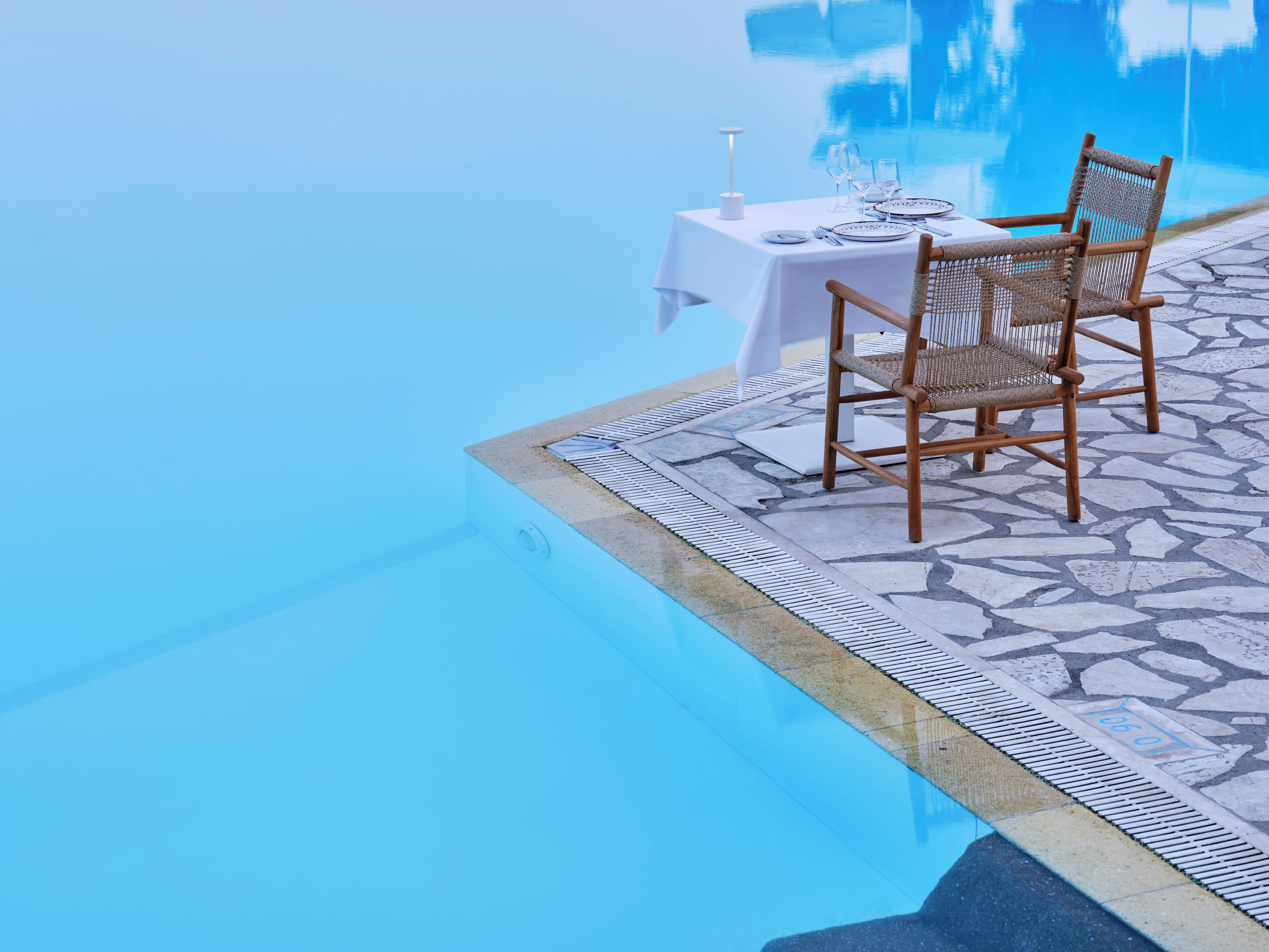 Mesa de comedor con sillas junto a una piscina, mantel blanco y una lámpara de mesa encendida.