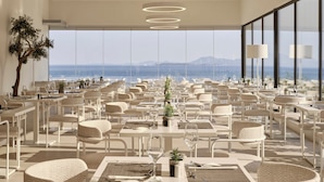 En modern restaurang med vita möbler, stora fönster med utsikt över havet och runda taklampor.