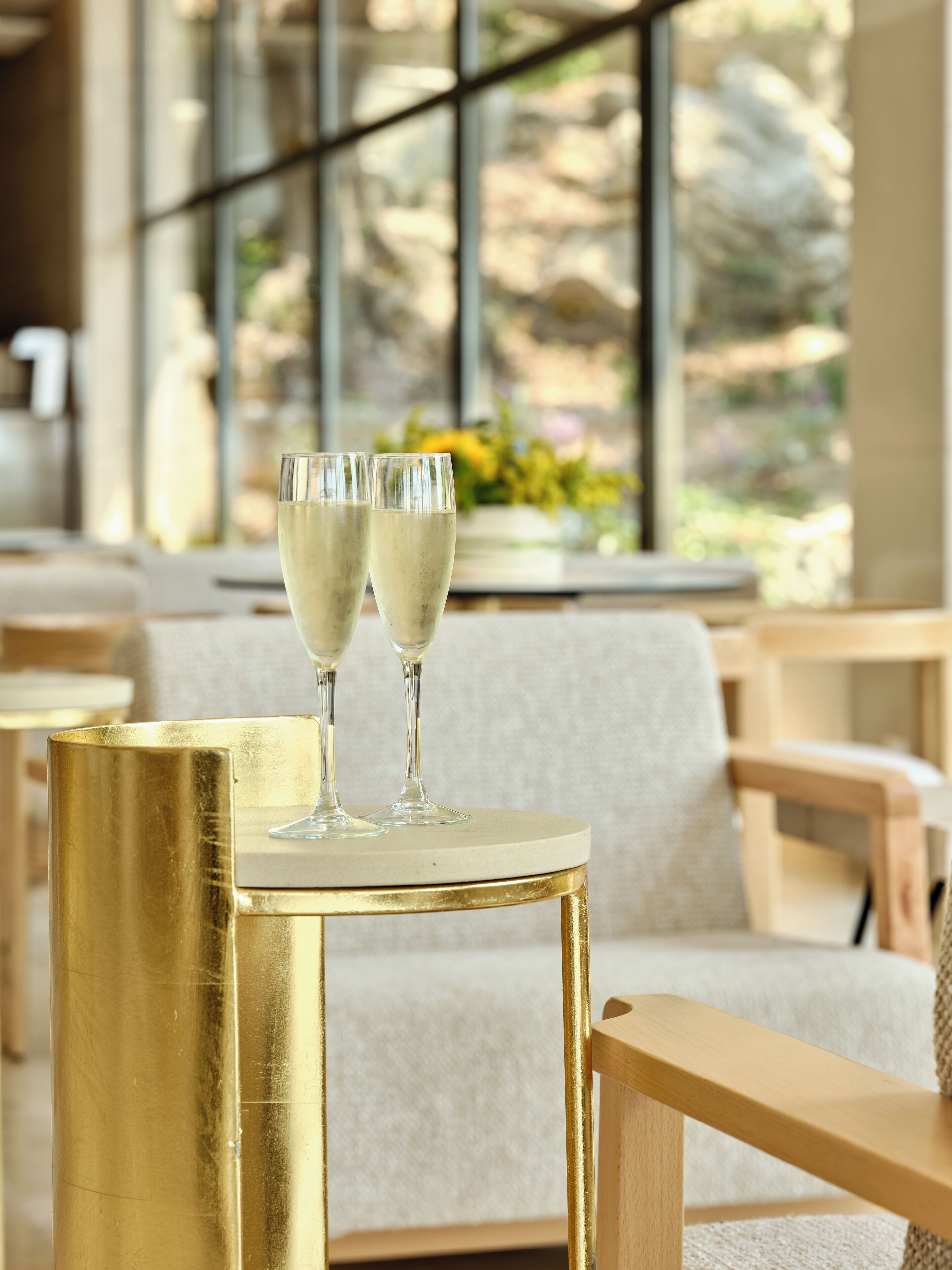 Two champagne flutes on a gold table in a well-lit room with a large window and a sofa.