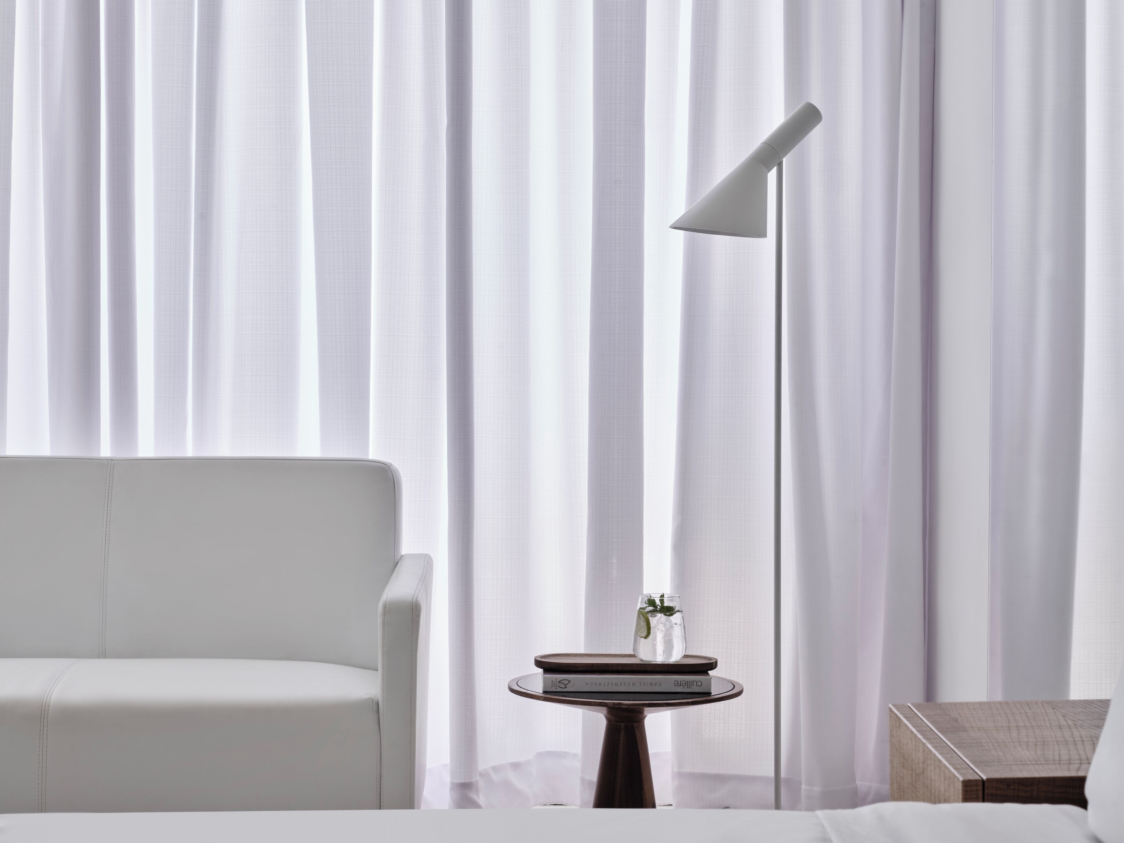 A modern hotel room with a white armchair, a floor lamp, a small table with a book and a glass of water, and sheer curtains.