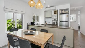 Una cocina moderna con una isla central, armarios blancos y un comedor con mesa y sillas de vidrio.