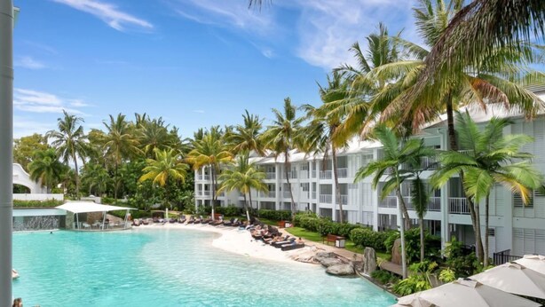 Un resort con una piscina grande rodeada de palmeras y un edificio blanco con balcones.
