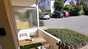 Een balkon met een tafel en stoelen, een kleine bloempot en uitzicht op een parkeerplaats en gebouwen.